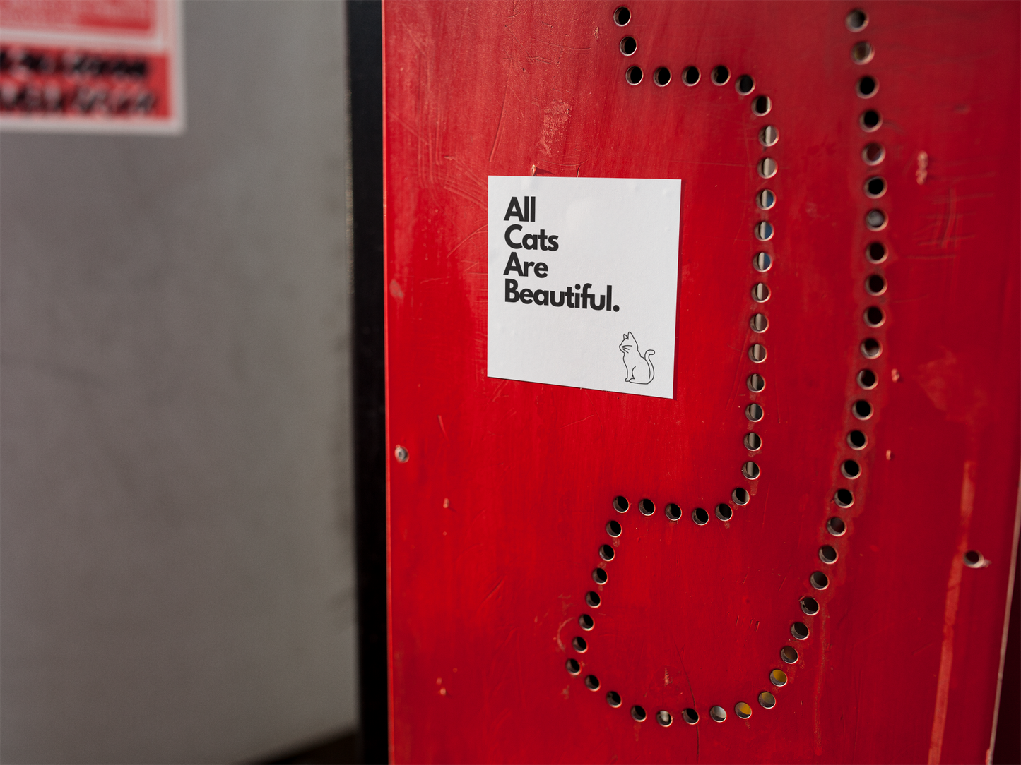 A white sticker featuring the bold statement 'all cats are bastards' in black block letters, complete with an outline of a cat below. Displayed on the side of a public red telephone booth